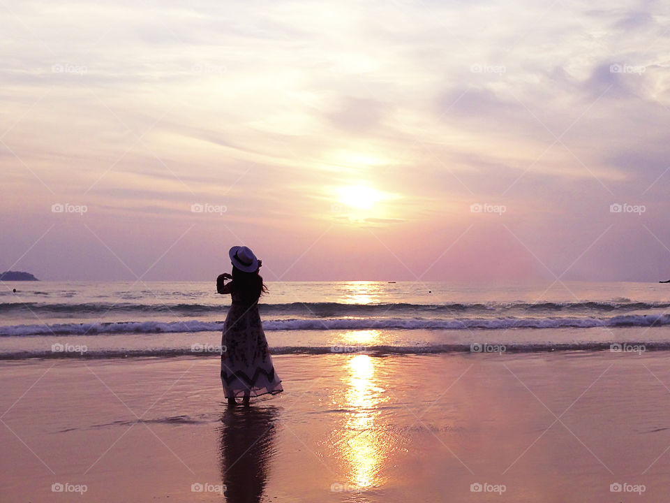 Silhouette of a young woman standing at the tropical sandy beach on sunset sky background 