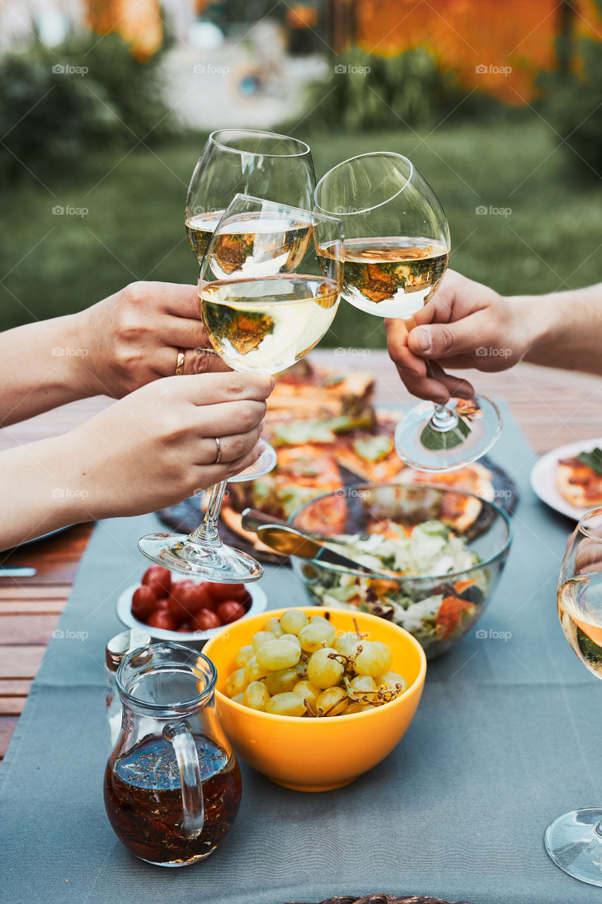 Friends making toast during summer picnic outdoor dinner in a home garden. Close up of people holding wine glasses with white wine over table with pizza, salads and fruits. Dinner in a orchard in a backyard
