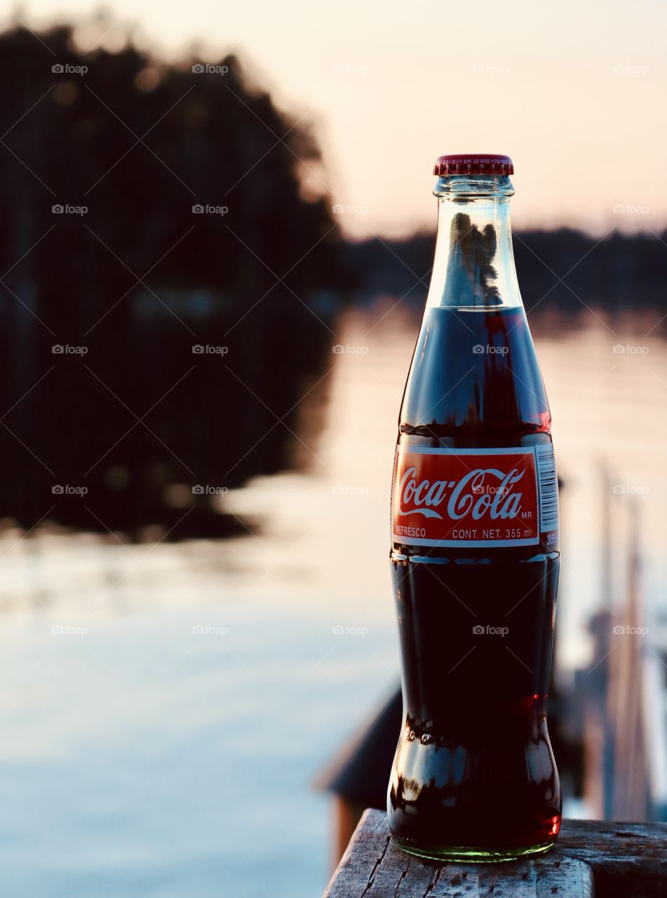 A traditional glass bottle of Coca Cola sits on the rail of a lakeside dock at sunset