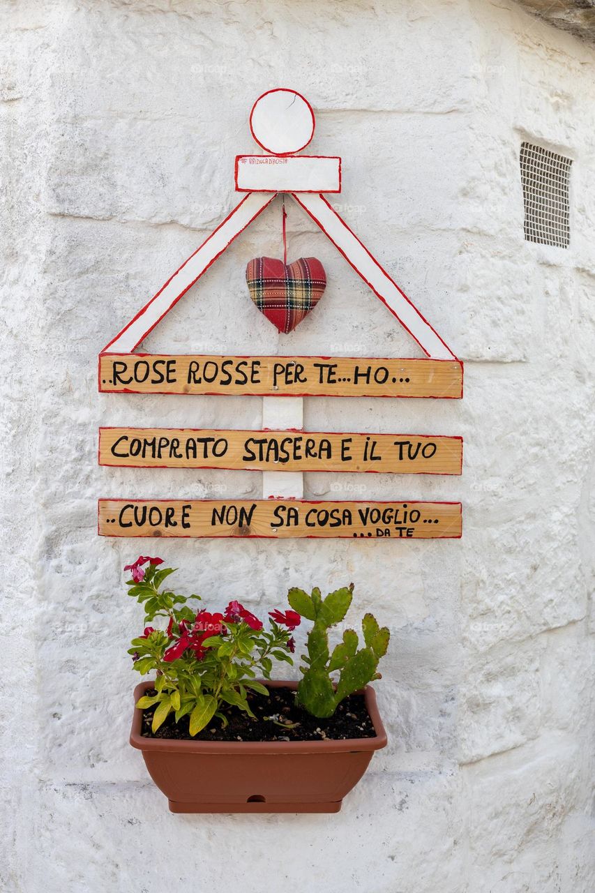 Beautiful side view of a flower pot and a wooden decoration with inscriptions hanging on the white stone wall of Trulla's house in the city of Alberabello in Italy, close-up side view. Concept urban plants.