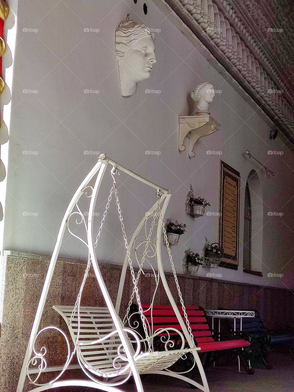 Urban art.  Place for rest.  White busts of ancient gods, pots of flowers hang on a white wall.  Near the wall are: a white swing bench, a red bench and a table
