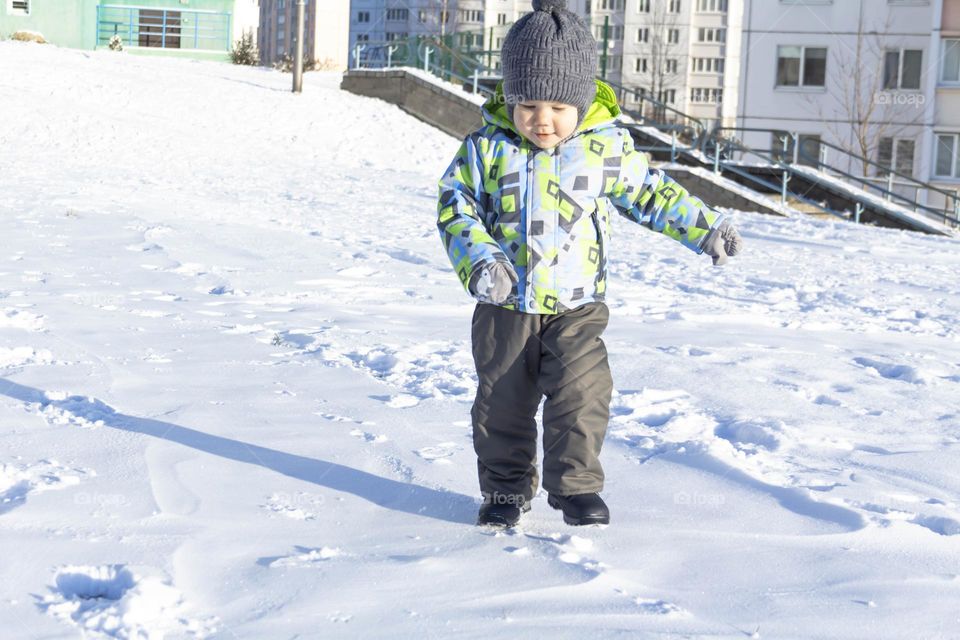 A child with a serious expression on his face in winter clothes jackets, pants, hat and boots in winter on the white snow on the street and in the park in nature plays winter fun.