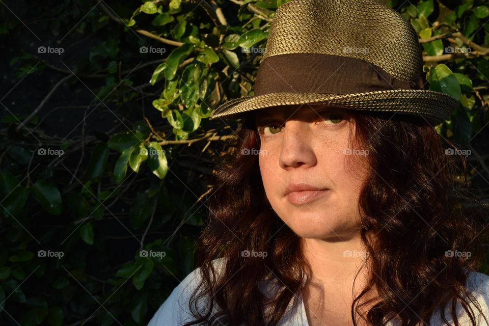 Women with natural and minimal makeup standing outdoors outside with wet hair. Women with red hair and green eyes standing next to tree taking a selfie. Sunlight shine on women hair and skin and making shadows. Women wearing a hat.