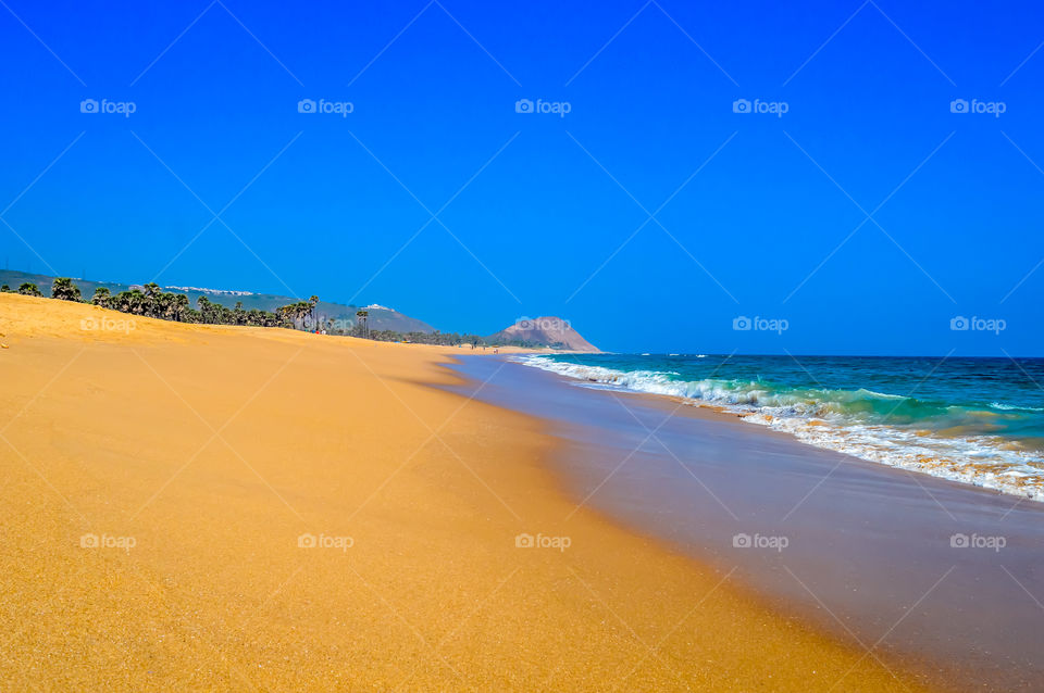 Wild Empty Tropical beach, vibrant yellow sand, bright blue sky, crystal clear waters with water crashing on the shore at daytime on a sunny day in in as landscape style may be used as a background, wallpaper, screen saver Travel vacation concept.