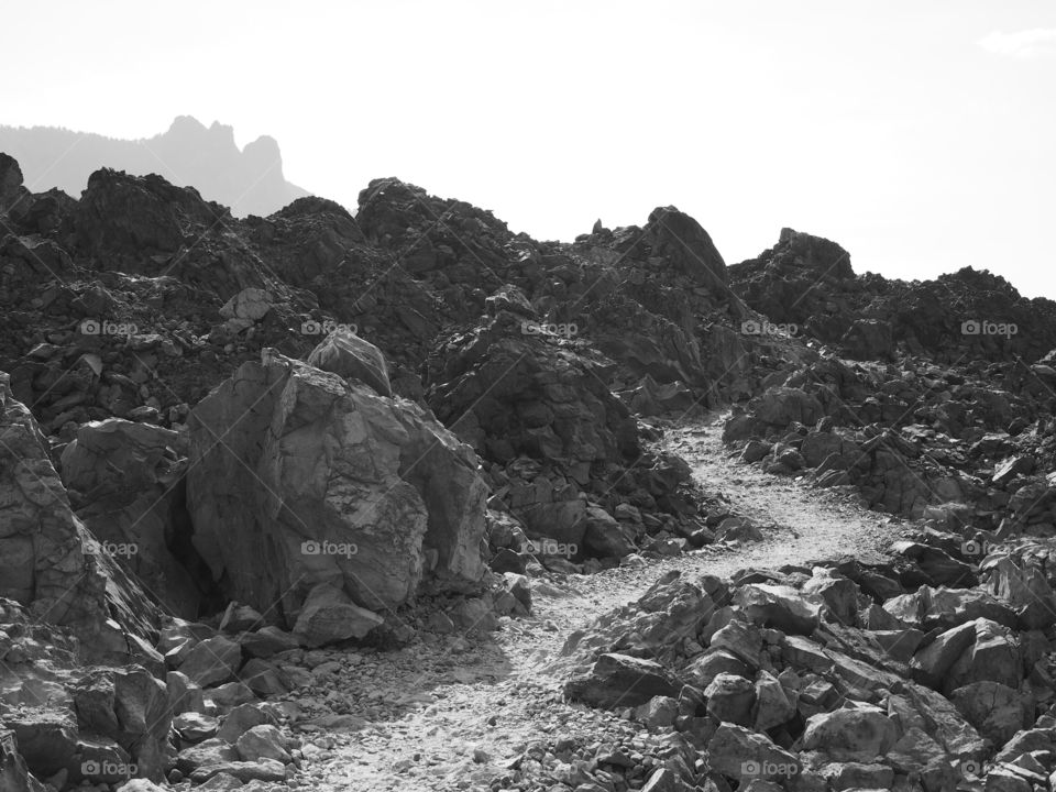 A rugged hiking trail winds through massive lava boulders with a mountain in the background at the Big Obsidian Flow in the Newberry National Volcanic Monument in Central Oregon on a fall day. 