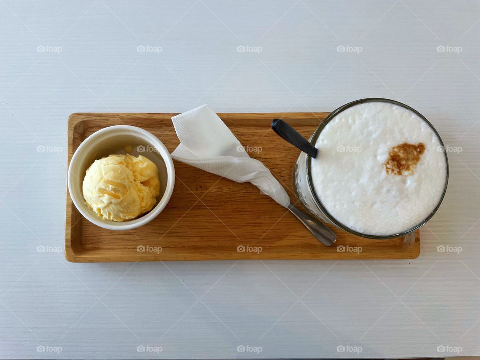 On top of a wooden tray are a fill latte, full ice cream cup, and a spoon wrapped in a tissue. They items are on a small wooden tray. 