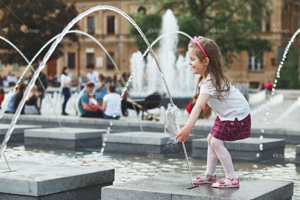 Little happy girl playing with the water in fountain in the center of town. Child catching a water stream going from a fountain. Candid people, real moments, authentic situations