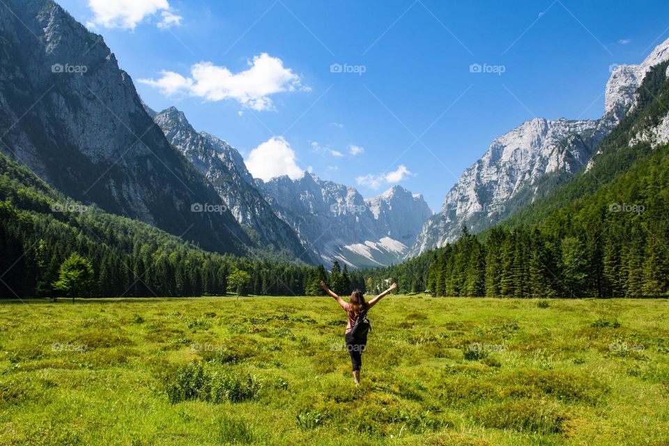 Rear view of young woman standing in the middle of a green valley surrounded by magnificent mountains