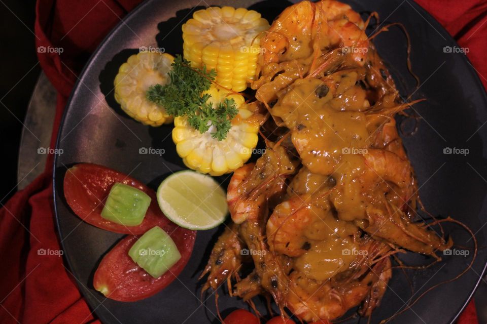 Asian food balado fried prawns with corn chunks, lemon slices and tomatoes served on black plate with red cloth bottom and wooden coaster. Asian style street seafood.