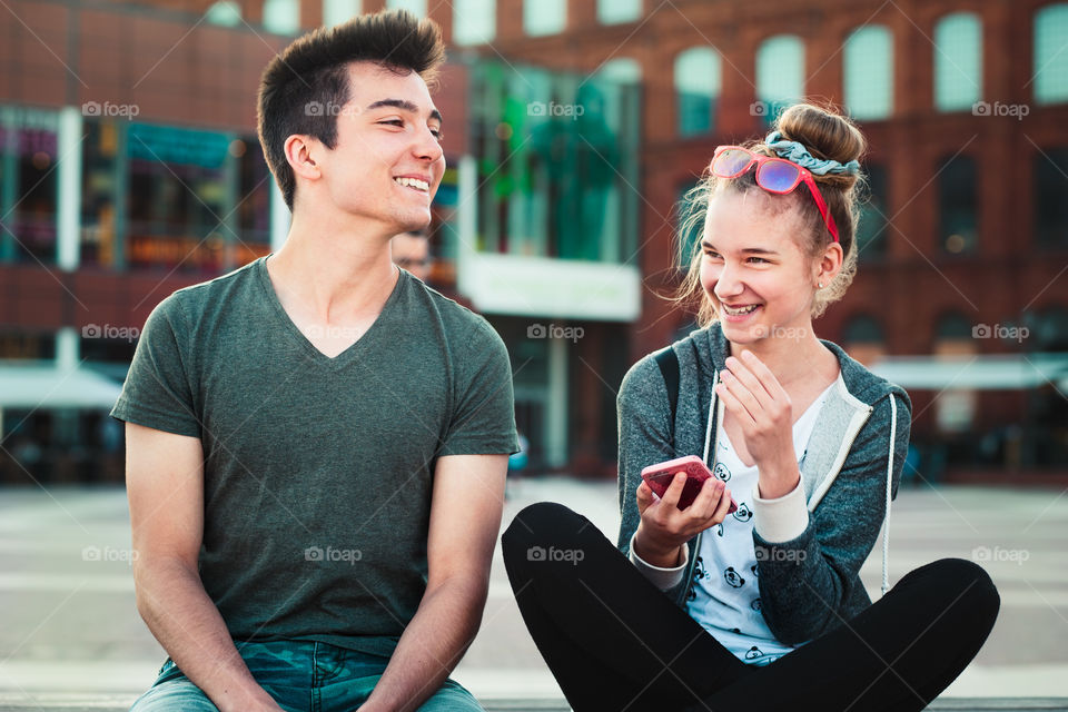 Couple of friends, teenage girl and boy, having fun together, using smartphones, sitting in center of town, spending time together. Real people, authentic situations