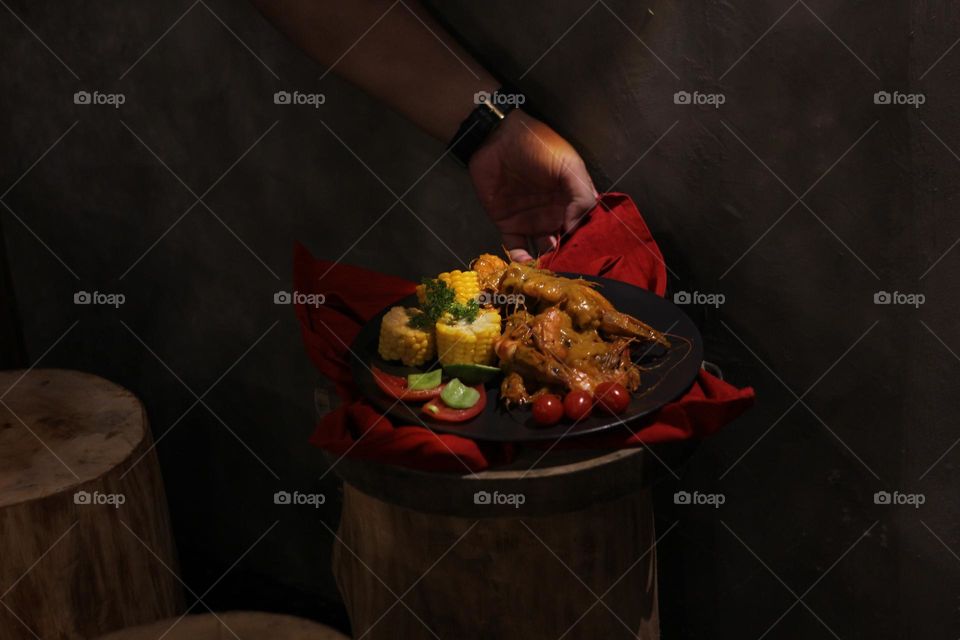 Asian food balado fried prawns with corn chunks, lemon slices and tomatoes served on black plate with red cloth bottom and wooden coaster. Asian style street seafood.