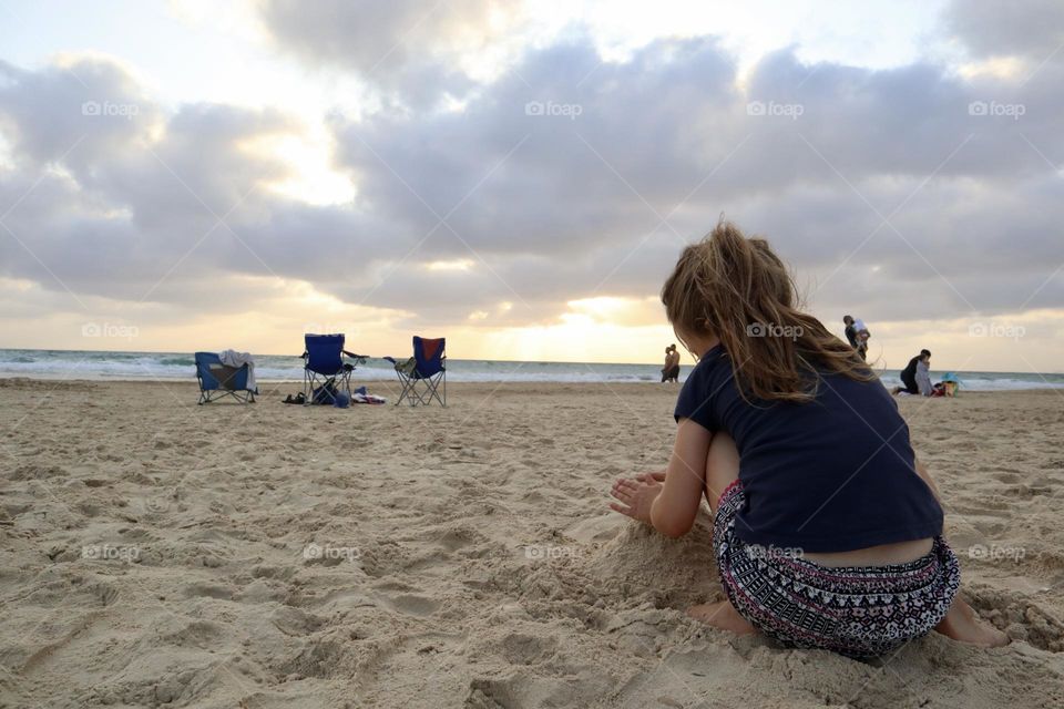 Girl playing in the sand on the beach during sunset with beach chairs on background 