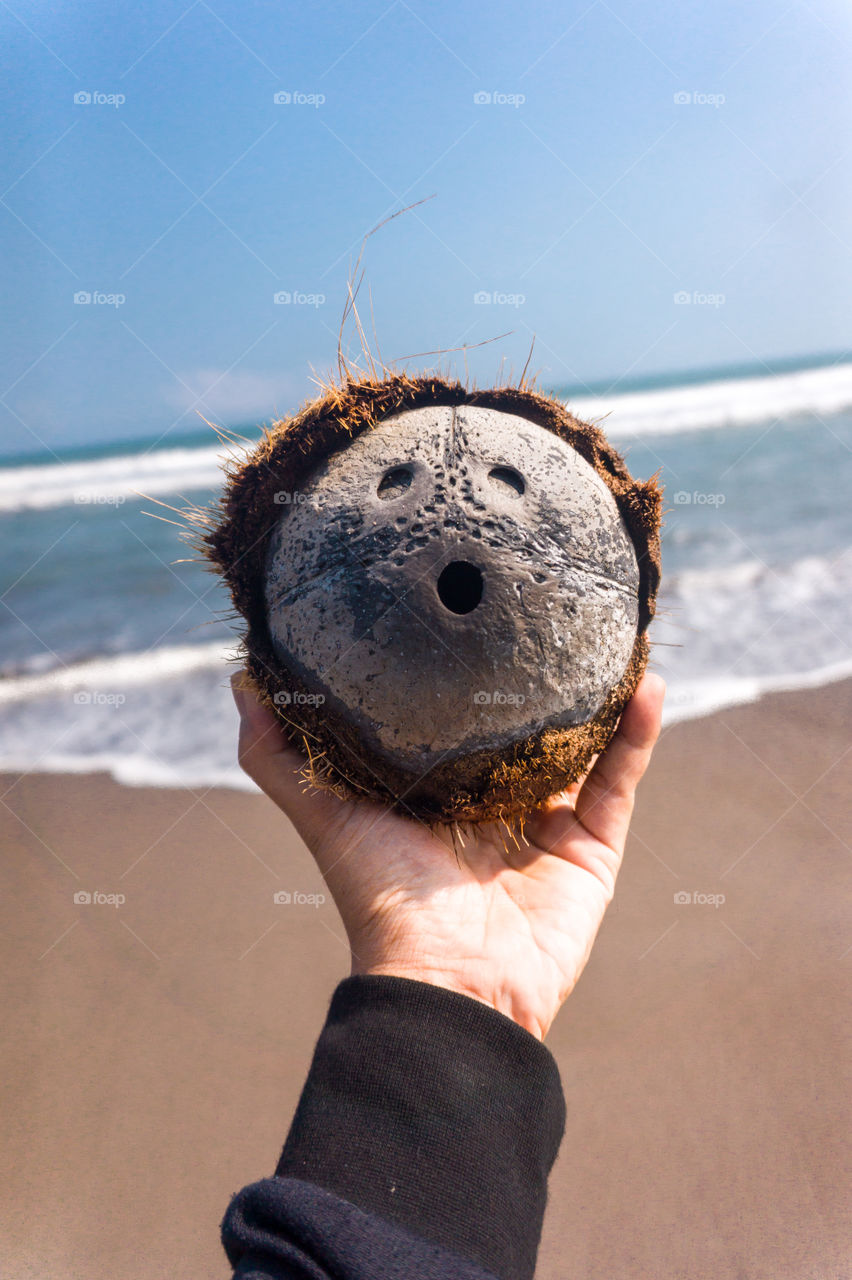 Close-up of a hand holding a coconut shell resembling a human face on an ocean background