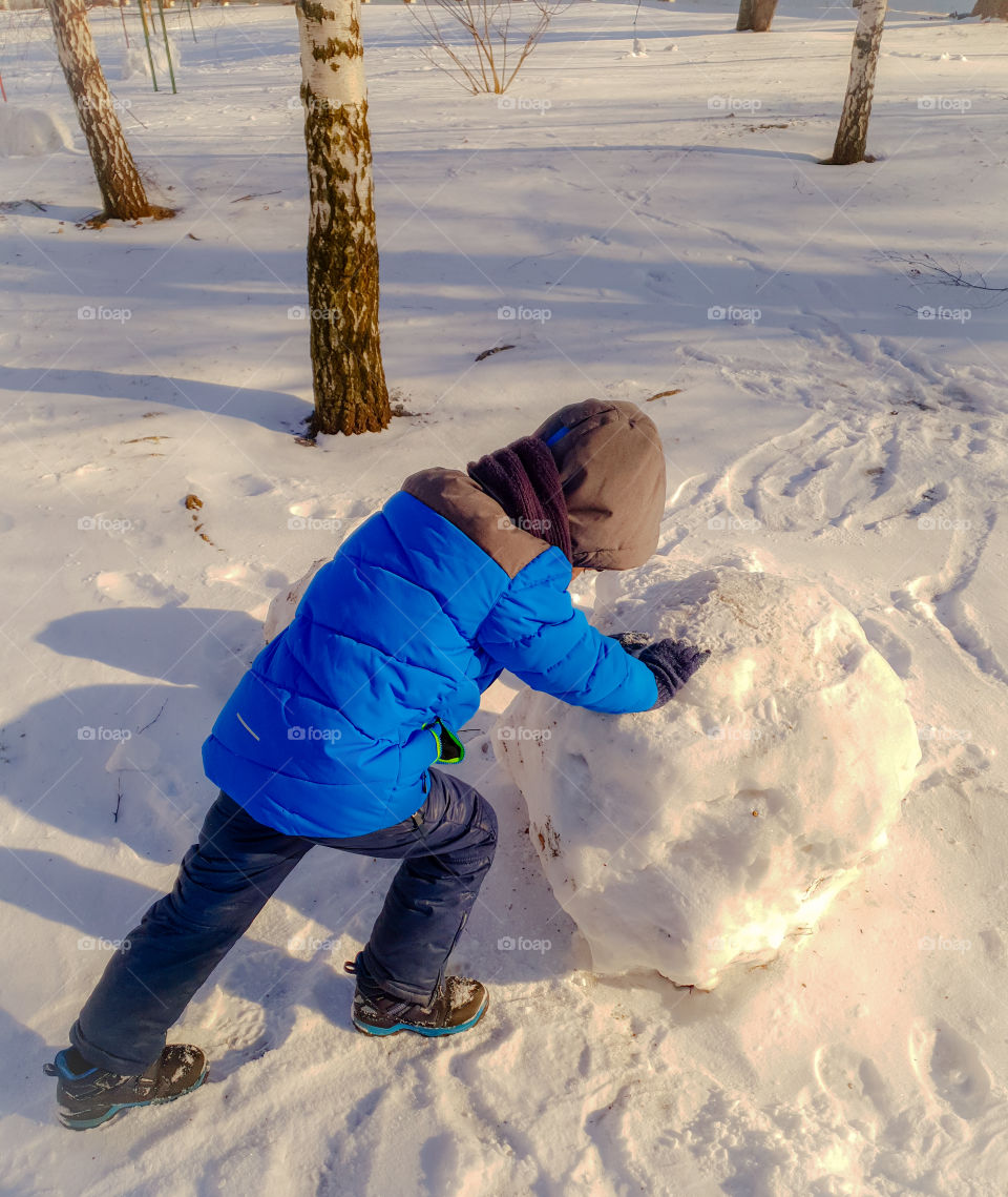 Winter sunny day in the park.  The boy rolls a snowball for a snowman.  Winter fun