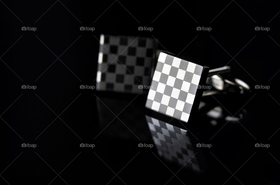 A close up portrait of two square cufflinks with a checkered pattern. they are surrounded by darkness and reflected onto a surface.