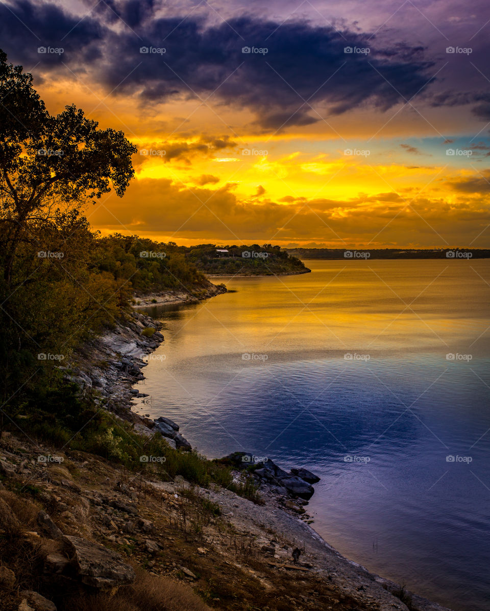 The sun sets over a rocky lake shoreline with a warm orange glow in the clouds