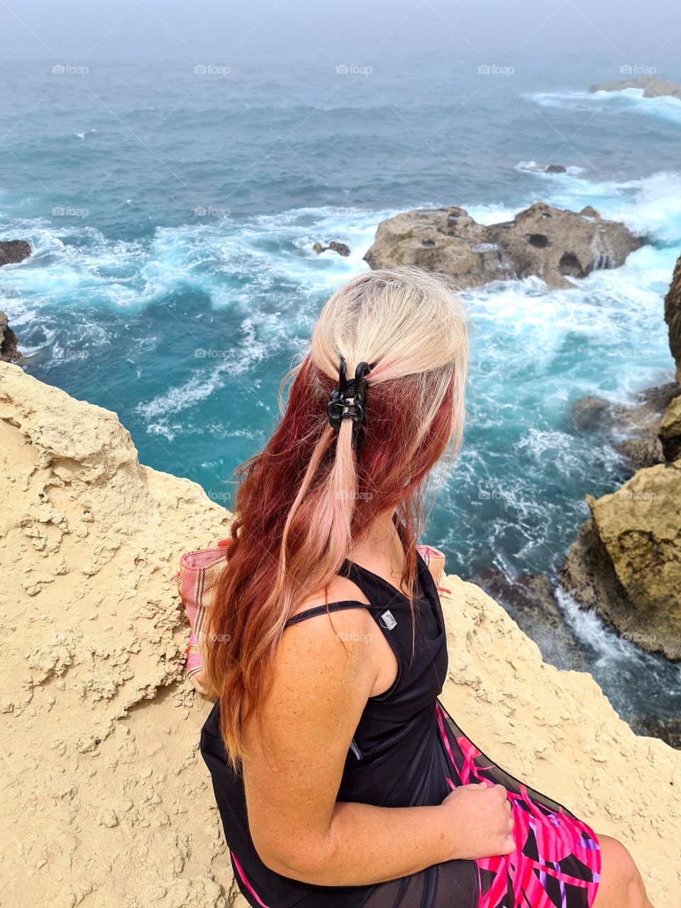 woman sitting on a rock cliff looking down at waves from the ocean crashing on the rocks below