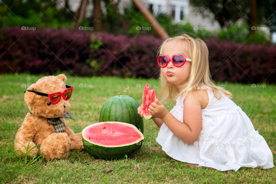 Cute little girl with blonde hair eating watermelon outdoor with stuffed teddy bear 