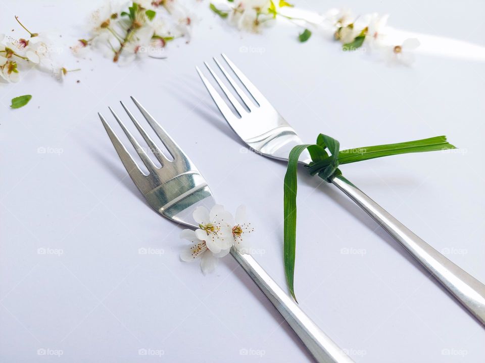 Two metal forks decorated with flowers and a grass tie,  as a woman and a man,  on a white table decorated with flowers