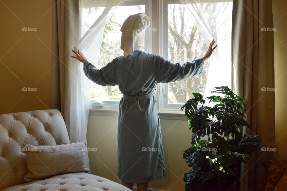 Back view of a woman wearing a robe and toweled hair opening the curtains of her bedroom window