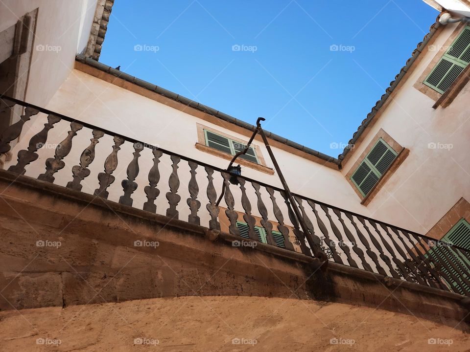View from the ground looking up of a courtyard at a Mediterranean traditional ancient Manor. Blue sky can be seen above. Facade of white walls, green Majorcan blinds and a metallic dark balustrade. Calvia, Majorca, Spain.