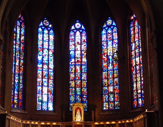 Very large and tall beautiful stained glass windows in a massive European cathedral provide bright colors and soft light on an alter. 