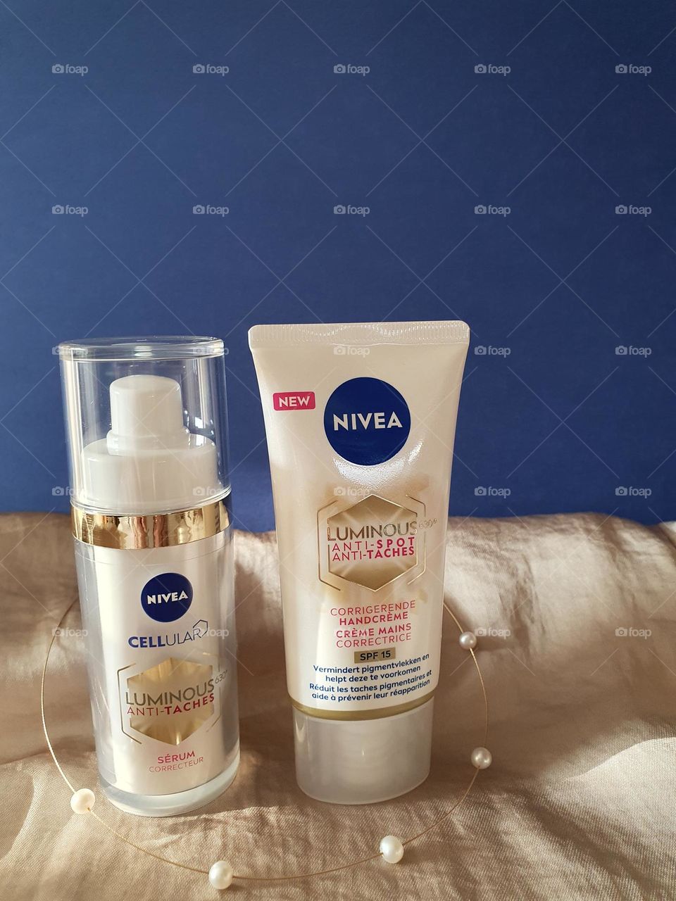 Nivea Cellular Luminous Anti-spot facial serum and handcream tubes presented on a golden satin fabric with a gold and pearls'necklace in front of a navy blue background