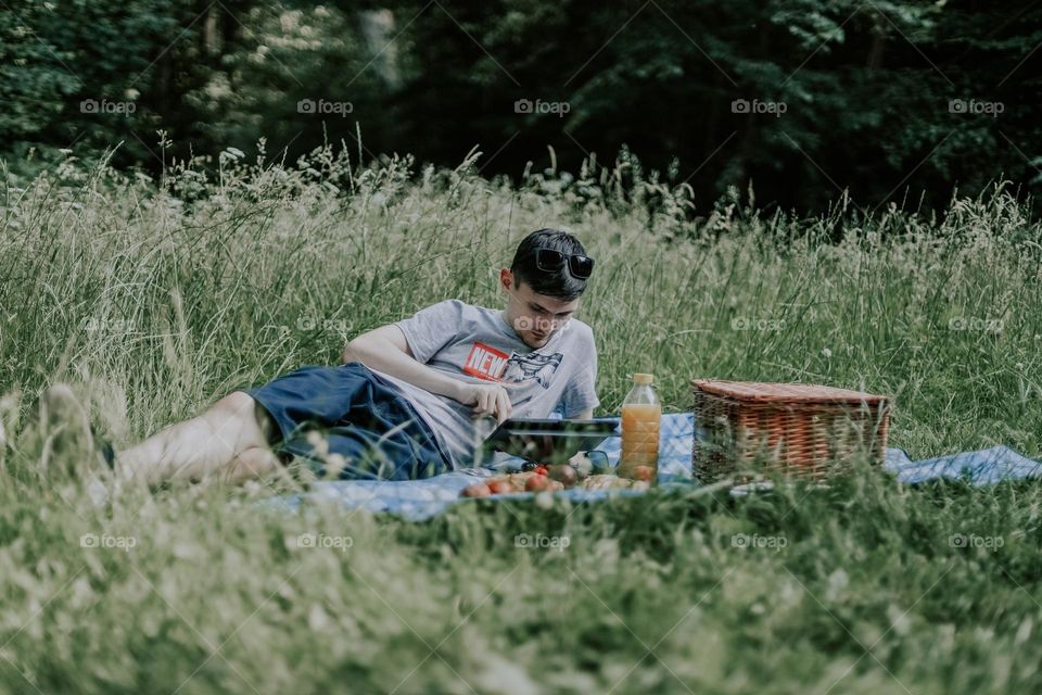 One young handsome caucasian brunette guy reclining on a bedspread with a wicker basket and fruits scans social networks in a tablet while relaxing in a meadow in a park in the summer sun in the afternoon, close-up side view. Male picnic time concept
