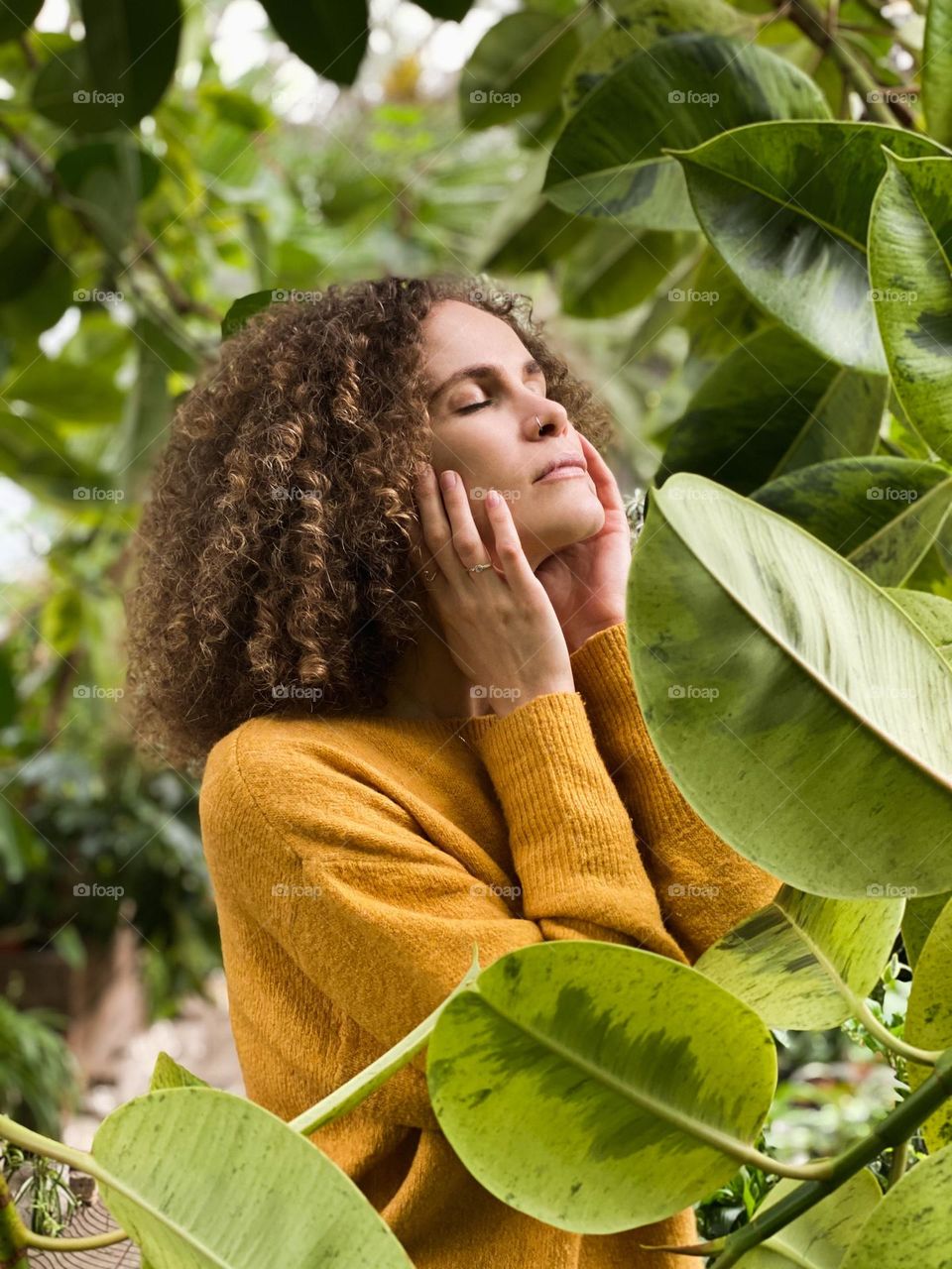 A young beautiful girl with curly hair in a yellow sweater stands with her eyes closed among the green plants in a botanical garden, portrait of woman 