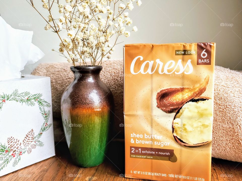 The new look Caress Six pack of shea butter and brown sugar bar soap next to a vase with flowers, a tissue box and a tan towel on a wood surface.