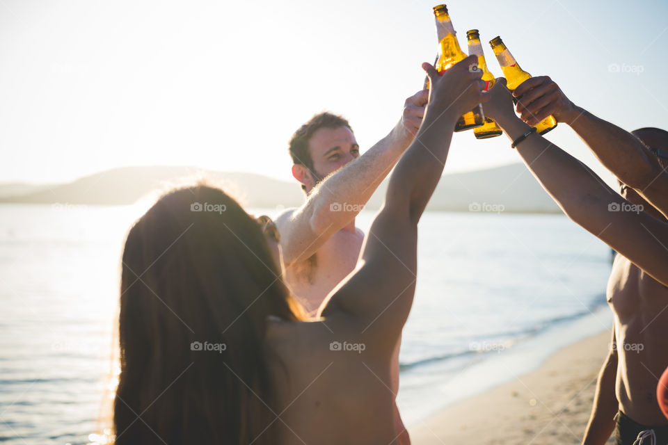 group of happy friends toasting with beer in the beach during sunset in summertime