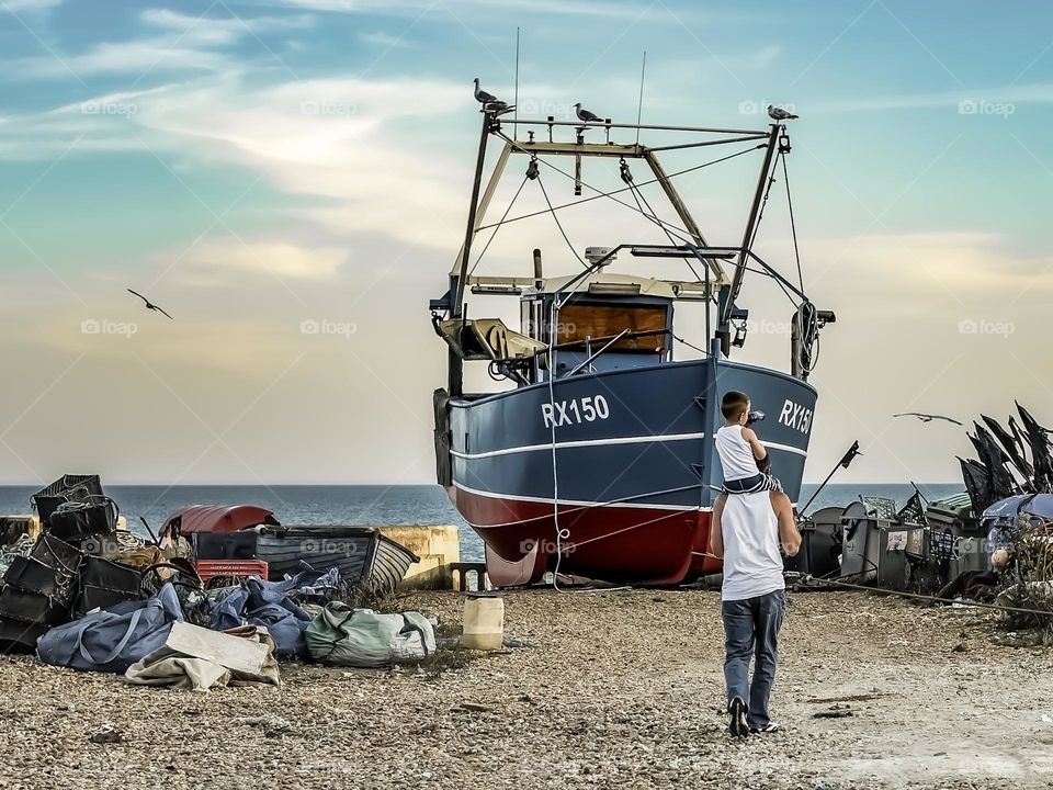 A man with a child on his shoulders, explore the working fishing beach at Hastings, UK in the late afternoon sunlight.