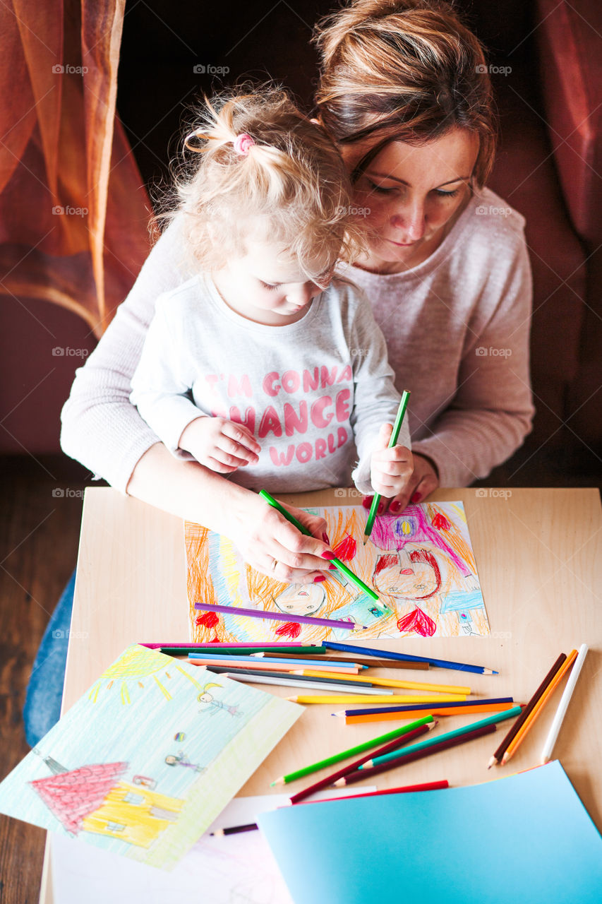 Mom with little daughter drawing a colorful pictures of family using pencil crayons sitting at table indoors. Shot from above