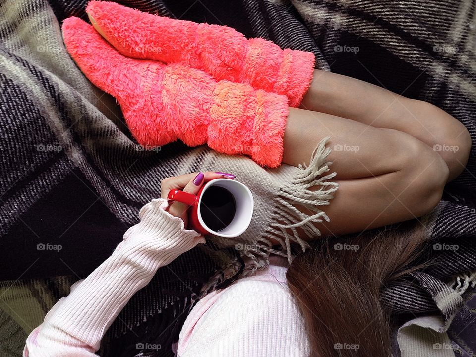 Young woman wearing cozy sweater and warm pink home boots enjoying hot coffee in cozy bed 