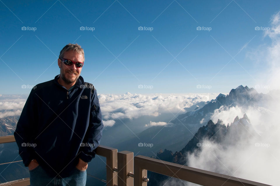 Bearded man with sunglasses stands before a vista of mountaintops