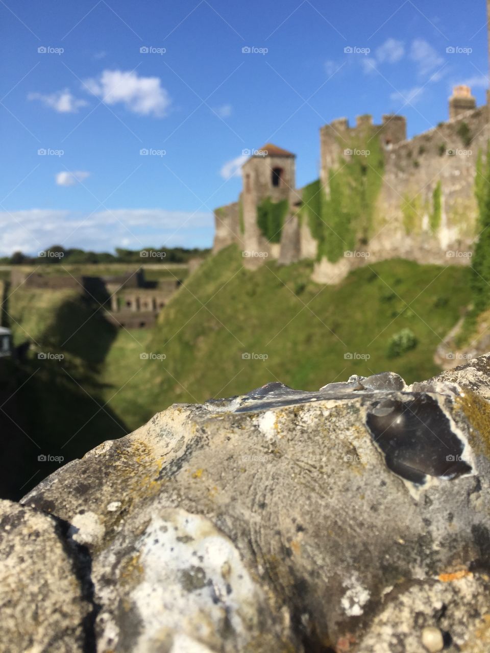 Castle views of vivid green fields, full of fresh grass and clear blue skies. Emerald green trees are lined up in rows in the horizon, with an intricate rock formation in the foreground 