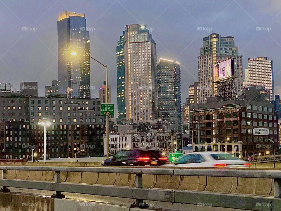 Coming home across the „Pulaski Bridge“ from „Bushwick“, Brooklyn at twilight time the last rays of the setting sun reflected in the high rises of „Long Island City“ with their nighttime illumination already turned on. 2023. Hypnotic Productions