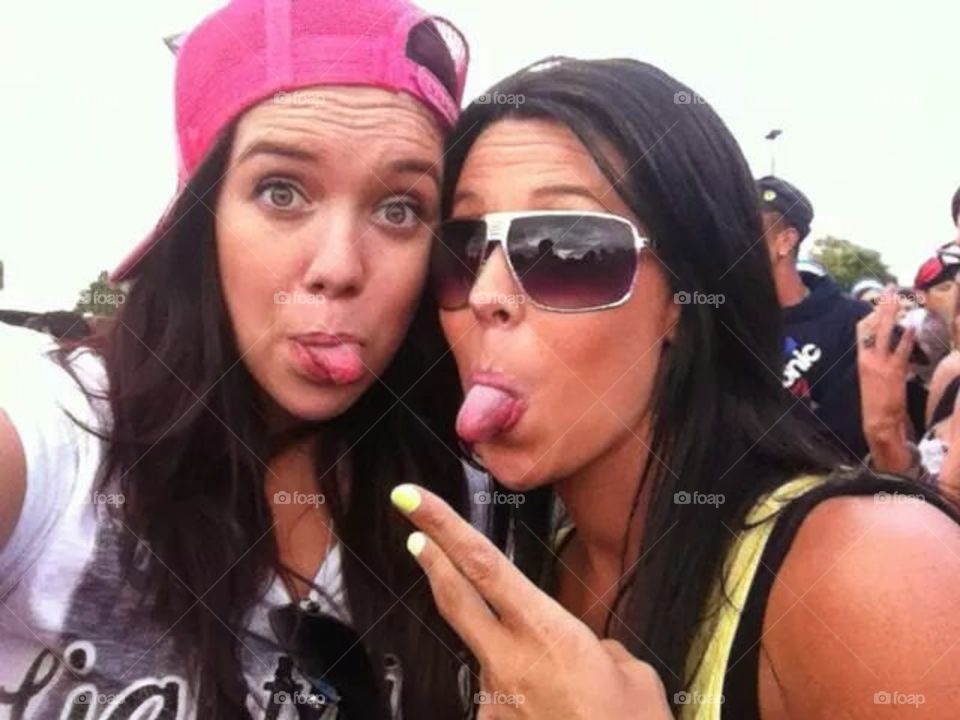 two women both poking there tongue out with one wearing sunglasses and one wearing a hat backwards at a music festival having fun