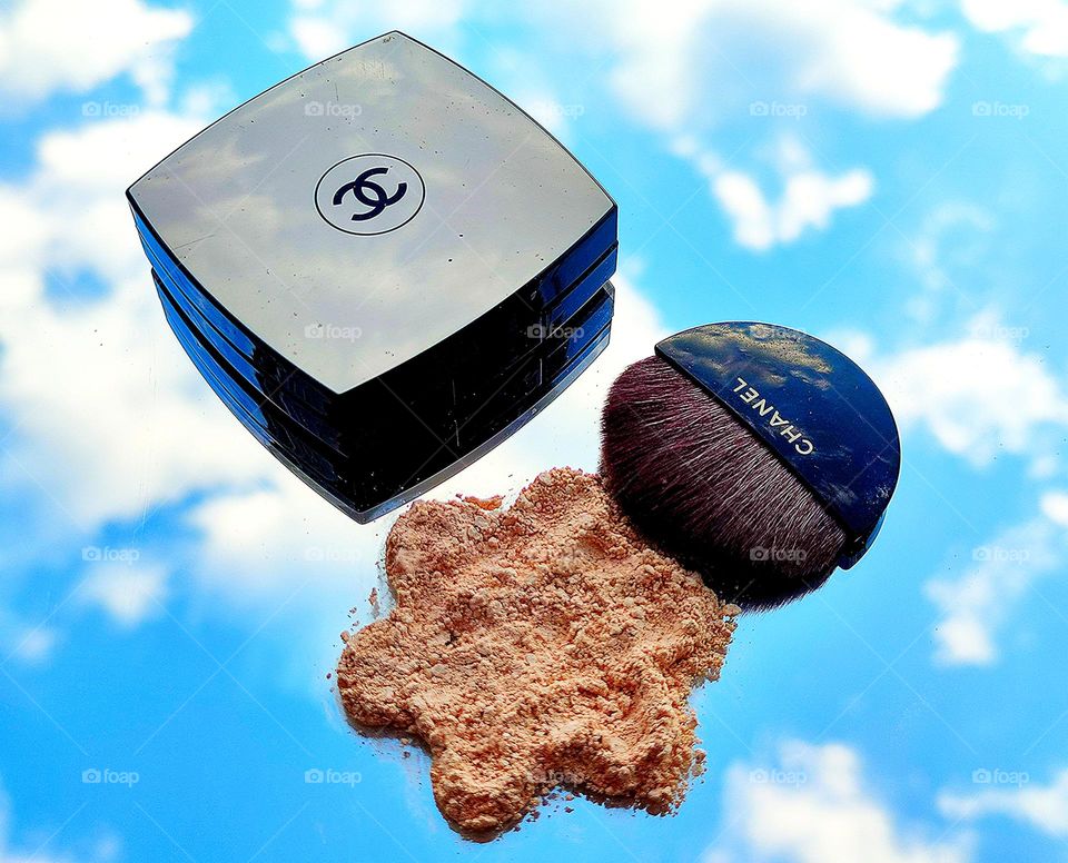 A box of powder with a logo, crushed pud in the shape of a flower and a black flat powder brush on a mirrored surface, which also reflects the blue sky with white clouds