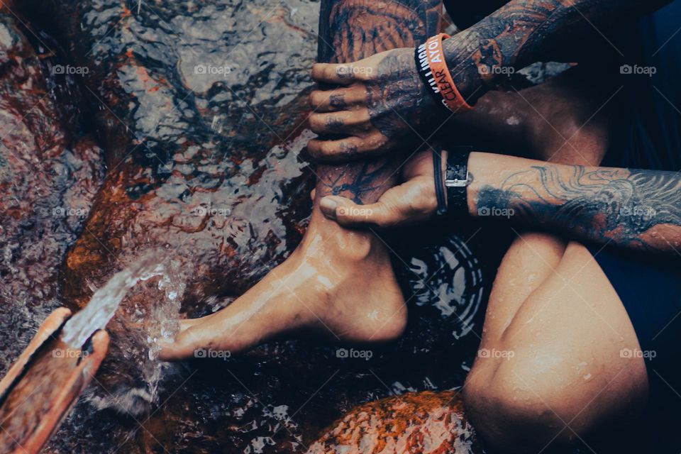 Under a bamboo shower, a tattooed man sits on a mossy rock holding his right leg, both hands and feet covered in tattoos.