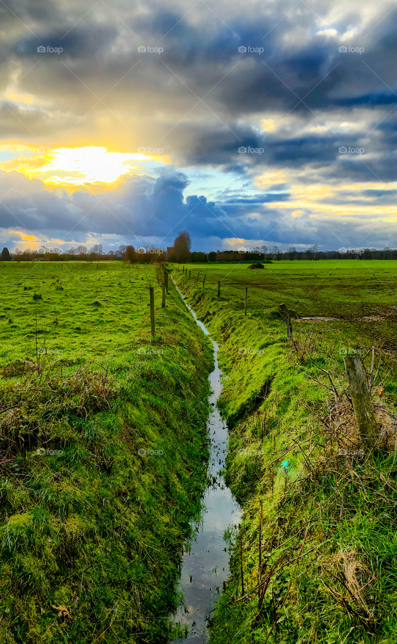 Golden sunrise or sunset sun shining through the dark stormy clouds over a rural countryside landscape showing grassy green farmfields divided by a creek