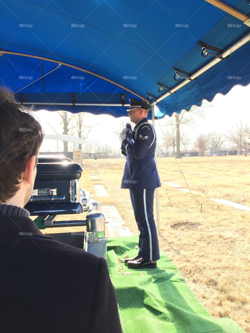 Military honor guard holding folded flag at graveside under blue awning with casket and family member witness