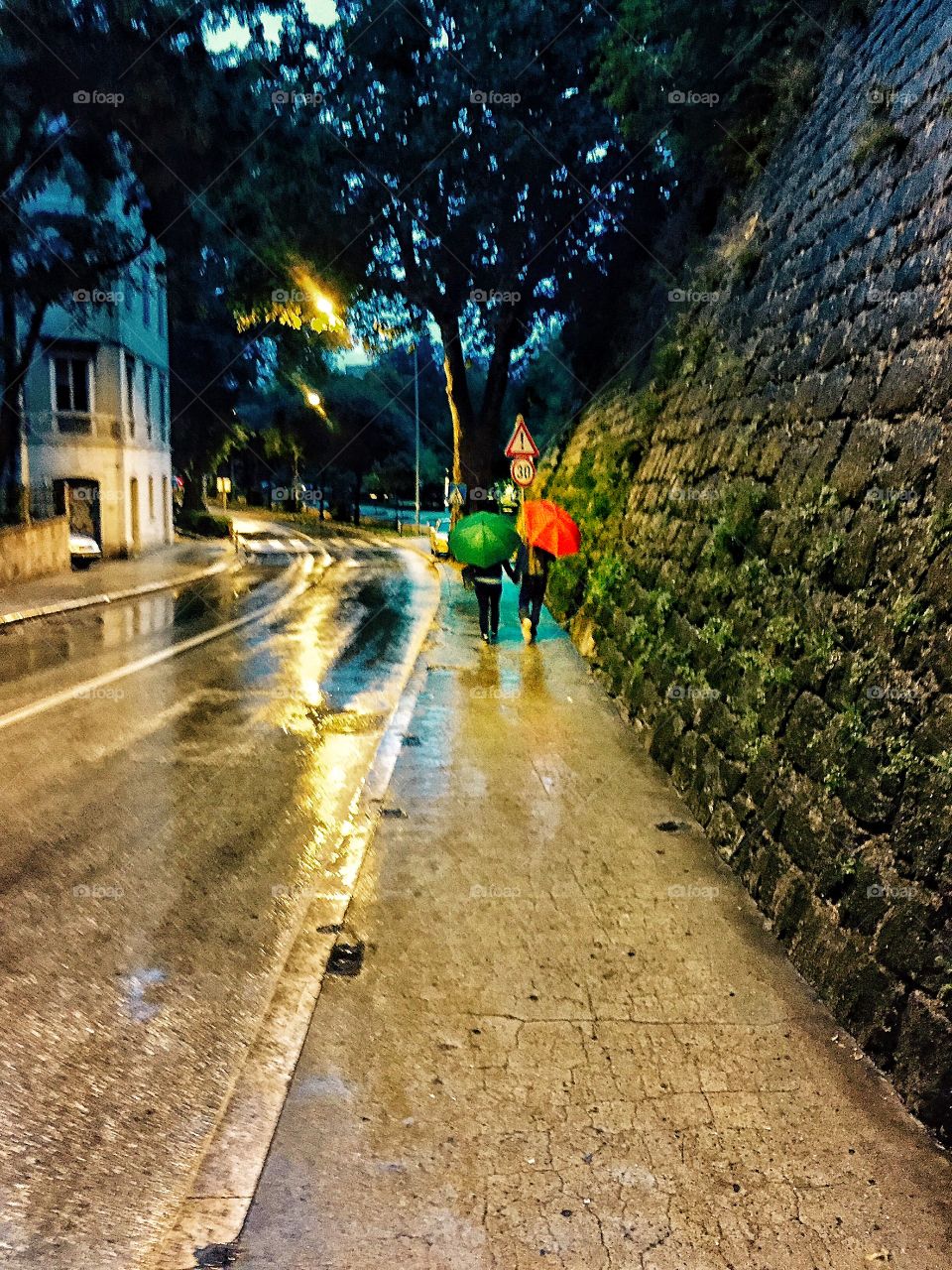 Couple walking in the rain with red and green umbrellas, city street in the evening