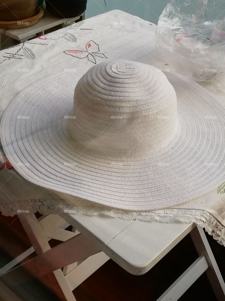 On a white stand patterned table cloth, slouch hat and an empty plastic box in a see-through wrinkled bag. A butterfly and green leaves embroidered on the textile. A lacy fabric around the cloth. A black flower pot and a pottery in the background.