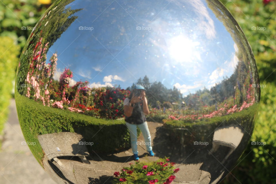 Woman with a camera capturing her image reflecting on a metal ball in the garden 