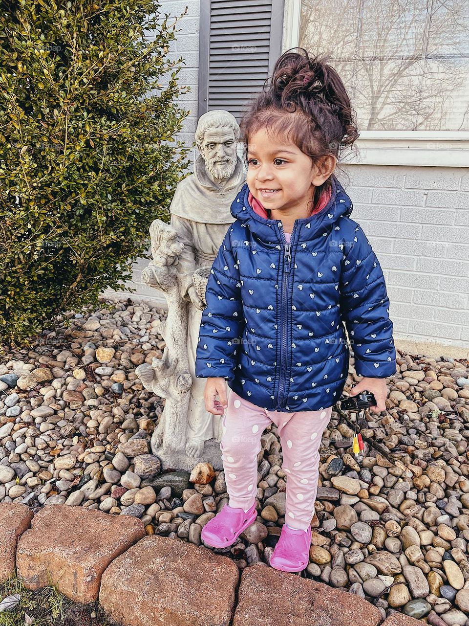 Toddler girl smiles with St. Francis statue, cement St. Francis Statue in front yard, little girl loves playing outside, interacting with statues, St. Francis decor, religious icons, St. Francis statue looking over toddler’s shoulder