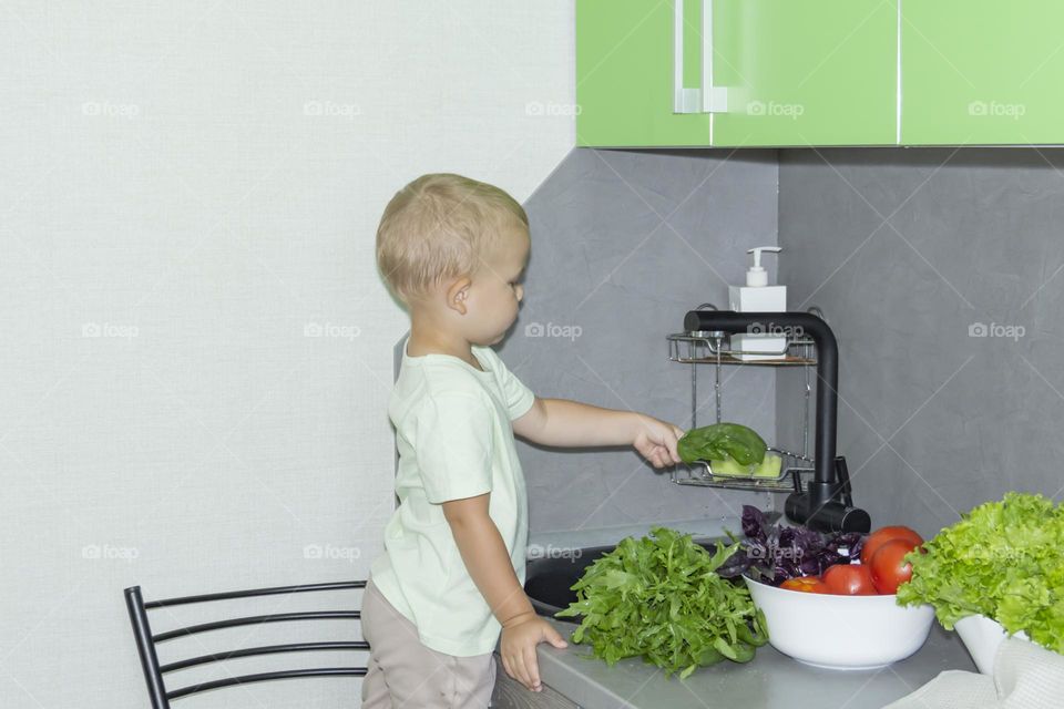 A small child washes fresh, green, vegetarian vegetables under the tap in a black sink to prepare salads and other dishes in a homemade gray and green kitchen.
