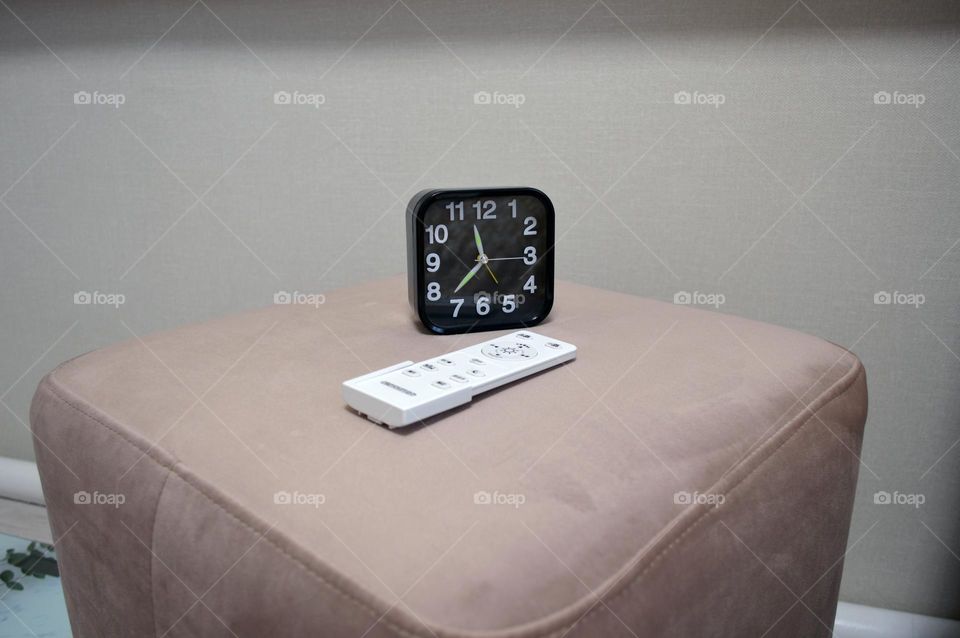 a black clock sits on the ottoman by the bed in the bedroom with a remote control