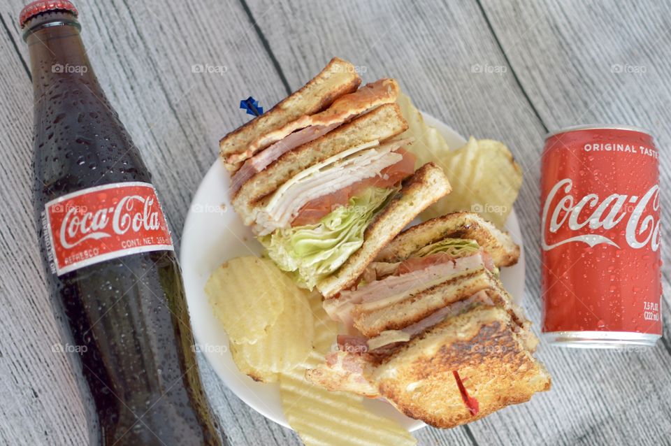 A can and a Bottle of Coca Cola with a club sandwich on a white rustic background 