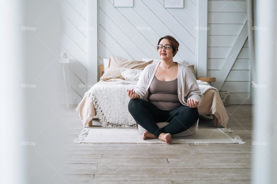 Middle aged plus size smiling brunette woman in home wear practicing yoga in bedroom
